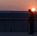 A silhouetted figure stands on an elevated flat surface, head bowed, holding a coiled cable or rope. The scene is backlit by a vivid sunset with a bright solar disk positioned just above the figure's head. A large industrial or hangar-style building with a corrugated metal roof is visible in the background.