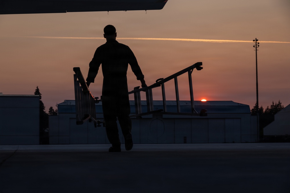 A silhouetted figure is captured on an airport tarmac during sunset or sunrise, carrying what appears to be a portable staircase or boarding ramp. The warm orange and red hues of the low sun contrast against the dark foreground, creating a dramatic silhouette composition. Large hangar buildings and tree lines are visible in the background.
