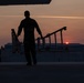 A silhouetted figure is captured on an airport tarmac during sunset or sunrise, carrying what appears to be a portable staircase or boarding ramp. The warm orange and red hues of the low sun contrast against the dark foreground, creating a dramatic silhouette composition. Large hangar buildings and tree lines are visible in the background.