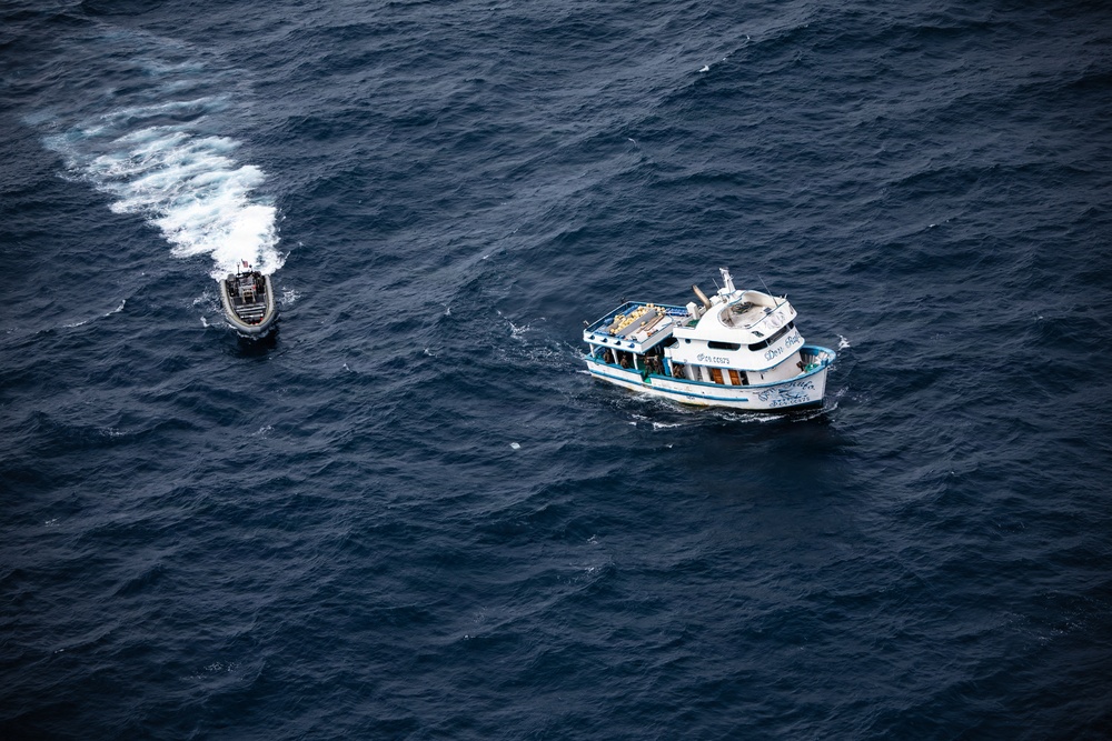 An aerial photograph shows two vessels on open dark blue ocean water. A larger white and blue civilian-style motor vessel is stationary or moving slowly on the right, while a smaller rigid inflatable boat (RIB) with a visible wake is approaching or circling it on the left. The scene suggests a maritime interception, patrol, or inspection operation.