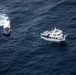 An aerial photograph shows two vessels on open dark blue ocean water. A larger white and blue civilian-style motor vessel is stationary or moving slowly on the right, while a smaller rigid inflatable boat (RIB) with a visible wake is approaching or circling it on the left. The scene suggests a maritime interception, patrol, or inspection operation.