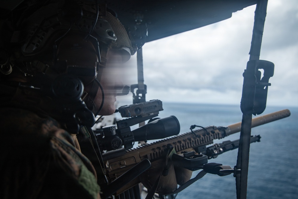 A military service member is positioned at the open door or ramp of an aircraft over open ocean, aiming a scoped rifle outward. The individual is wearing full tactical gear including a combat helmet with communications equipment. The image appears to depict aerial overwatch or a door-gunner/sniper training or operational scenario.