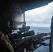 A military service member is positioned at the open door or ramp of an aircraft over open ocean, aiming a scoped rifle outward. The individual is wearing full tactical gear including a combat helmet with communications equipment. The image appears to depict aerial overwatch or a door-gunner/sniper training or operational scenario.