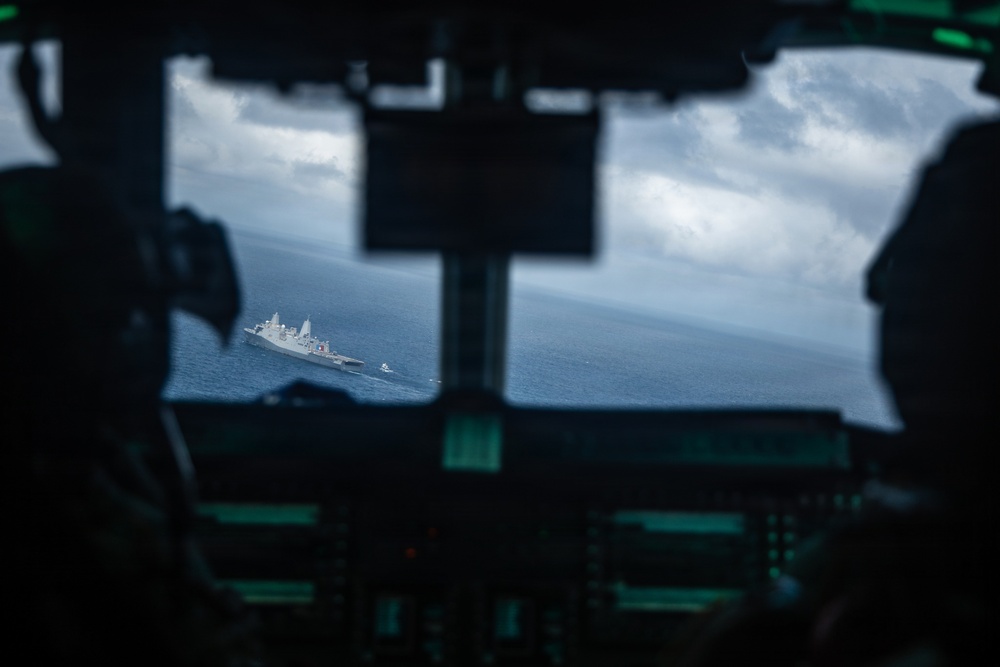 The image is taken from inside a military aircraft cockpit, looking through the windshield at a naval warship sailing on open ocean below. Two crew members are partially visible in silhouette on either side of the frame, with cockpit instrumentation visible in the foreground. The scene suggests an aerial surveillance or coordination mission between air and naval assets.