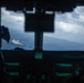 The image is taken from inside a military aircraft cockpit, looking through the windshield at a naval warship sailing on open ocean below. Two crew members are partially visible in silhouette on either side of the frame, with cockpit instrumentation visible in the foreground. The scene suggests an aerial surveillance or coordination mission between air and naval assets.