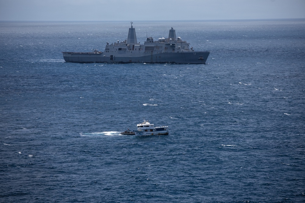 The image shows a large U.S. Navy amphibious transport dock ship bearing hull number 17 anchored or underway in open blue ocean waters. In the foreground, a smaller civilian-style white ferry or patrol vessel is moving through the water, accompanied by what appears to be a small rigid inflatable boat (RIB). The scene suggests a maritime interaction or boarding operation between naval and civilian vessels.