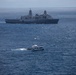 The image shows a large U.S. Navy amphibious transport dock ship bearing hull number 17 anchored or underway in open blue ocean waters. In the foreground, a smaller civilian-style white ferry or patrol vessel is moving through the water, accompanied by what appears to be a small rigid inflatable boat (RIB). The scene suggests a maritime interaction or boarding operation between naval and civilian vessels.