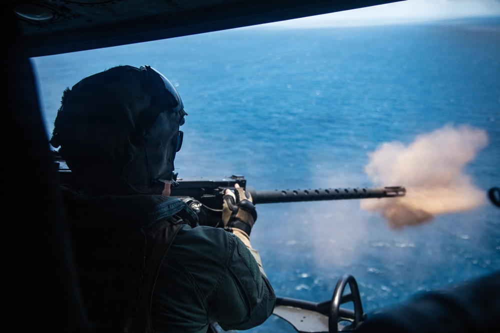 A military aircrew member is firing a door-mounted machine gun from a helicopter over open ocean. The weapon is actively discharging, with a visible muzzle flash and smoke cloud at the barrel. The shooter is wearing full flight gear including a helmet, flight suit, and tactical gloves.