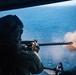 A military aircrew member is firing a door-mounted machine gun from a helicopter over open ocean. The weapon is actively discharging, with a visible muzzle flash and smoke cloud at the barrel. The shooter is wearing full flight gear including a helmet, flight suit, and tactical gloves.