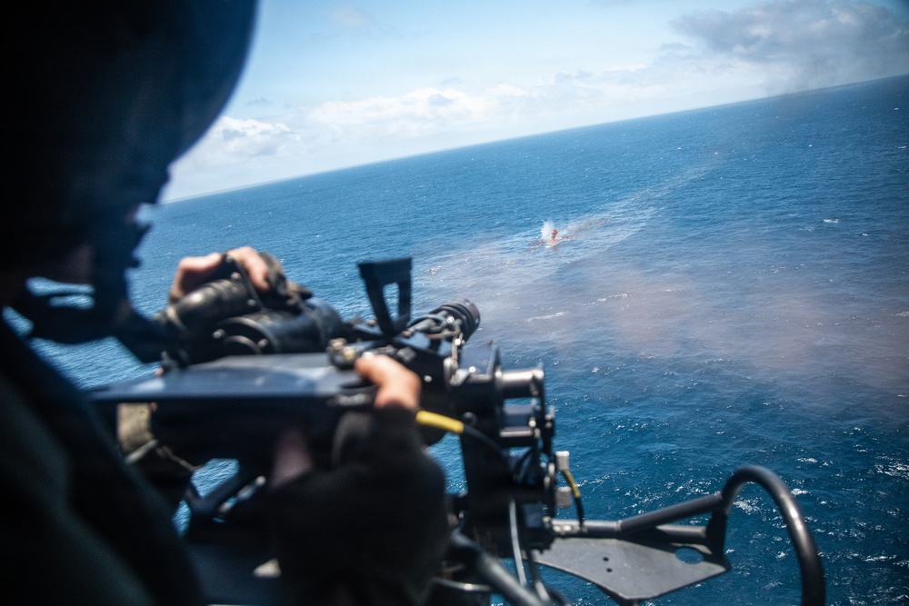A military personnel is operating a door-mounted machine gun from an airborne helicopter over open ocean. Rounds are visibly impacting the water surface near what appears to be a target or marker in the distance. This appears to be a live-fire training exercise conducted from a rotary-wing aircraft.