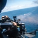 A military personnel is operating a door-mounted machine gun from an airborne helicopter over open ocean. Rounds are visibly impacting the water surface near what appears to be a target or marker in the distance. This appears to be a live-fire training exercise conducted from a rotary-wing aircraft.