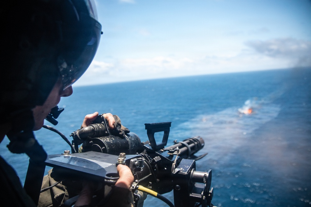 A military aircrew member wearing a flight helmet is operating a door-mounted machine gun from a helicopter over open ocean. Smoke or a flare is visible on the water surface in the background, suggesting an active firing exercise or live-fire training event. The perspective indicates the photo was taken from inside the helicopter looking outward.