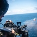 A military aircrew member wearing a flight helmet is operating a door-mounted machine gun from a helicopter over open ocean. Smoke or a flare is visible on the water surface in the background, suggesting an active firing exercise or live-fire training event. The perspective indicates the photo was taken from inside the helicopter looking outward.
