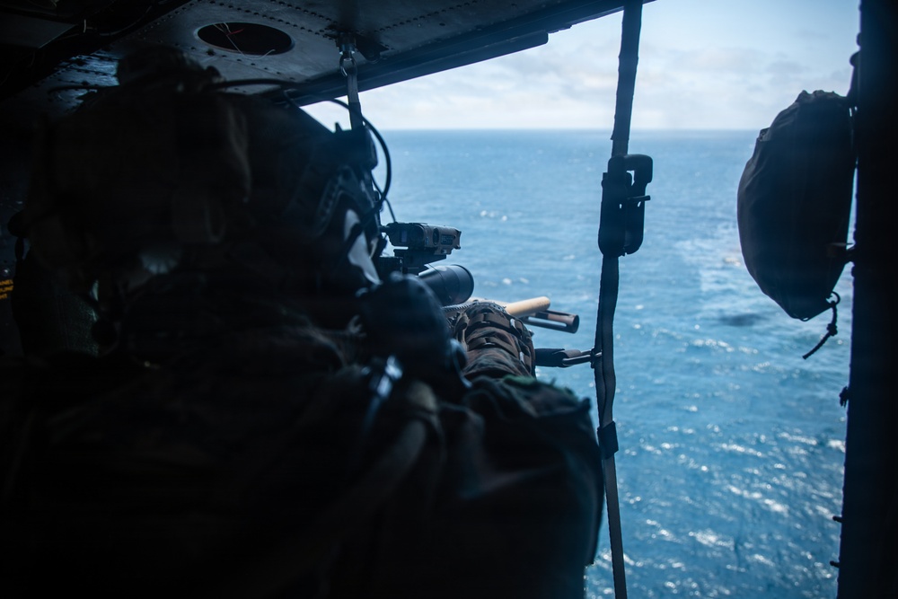 A military personnel member is positioned inside an open helicopter door, aiming a scoped rifle toward the ocean below. The shooter is wearing full tactical gear including a helmet with communications equipment. A second helmeted individual is partially visible on the right side of the frame.