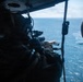 A military personnel member is positioned inside an open helicopter door, aiming a scoped rifle toward the ocean below. The shooter is wearing full tactical gear including a helmet with communications equipment. A second helmeted individual is partially visible on the right side of the frame.
