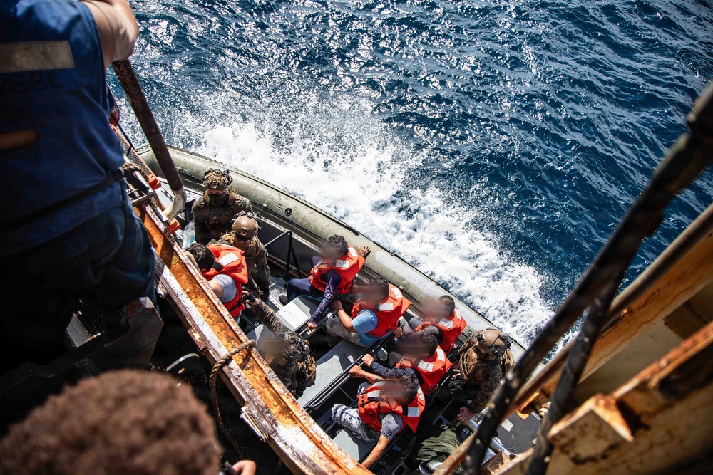 The image shows a maritime rescue or boarding operation at sea, with multiple individuals wearing orange life vests seated in a rigid inflatable boat (RIB) alongside what appears to be a larger vessel. Armed personnel in tactical gear are present on the boat, suggesting a military or coast guard operation. The faces of several individuals have been blurred, indicating editorial redaction for privacy or security purposes.