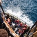 The image shows a maritime rescue or boarding operation at sea, with multiple individuals wearing orange life vests seated in a rigid inflatable boat (RIB) alongside what appears to be a larger vessel. Armed personnel in tactical gear are present on the boat, suggesting a military or coast guard operation. The faces of several individuals have been blurred, indicating editorial redaction for privacy or security purposes.