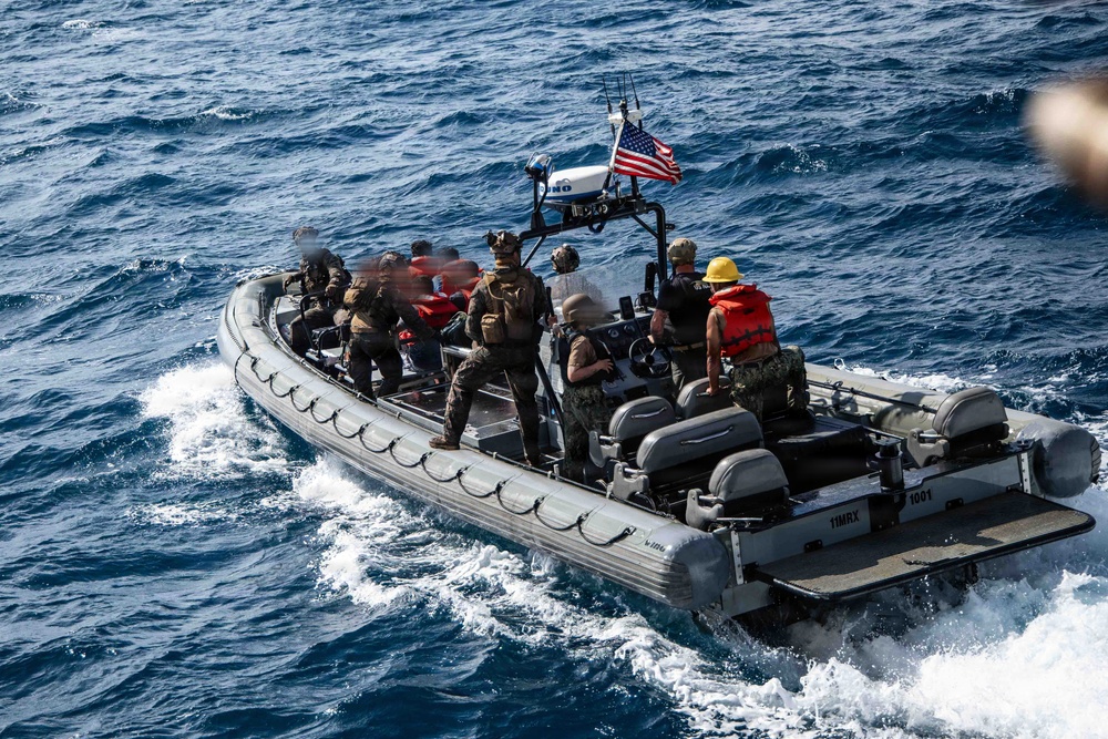 A rigid-hull inflatable boat (RHIB) marked '11MRX 1001' is underway on open ocean waters carrying multiple military personnel. The boat flies a United States flag and is equipped with a mast-mounted sensor or camera system. Personnel aboard are wearing a mix of tactical gear, life vests, and helmets, suggesting a military training or operational mission.