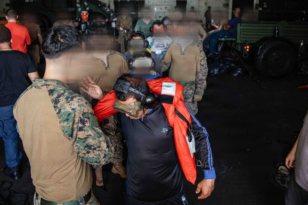The image shows a group of military personnel and civilians inside what appears to be a naval vessel's hangar or well deck. Several individuals in U.S. Marine Corps MARPAT camouflage uniforms are present alongside civilians wearing orange life vests and hearing protection headsets. The scene suggests an evacuation, rescue, or embarkation operation.