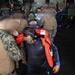 The image shows a group of military personnel and civilians inside what appears to be a naval vessel's hangar or well deck. Several individuals in U.S. Marine Corps MARPAT camouflage uniforms are present alongside civilians wearing orange life vests and hearing protection headsets. The scene suggests an evacuation, rescue, or embarkation operation.