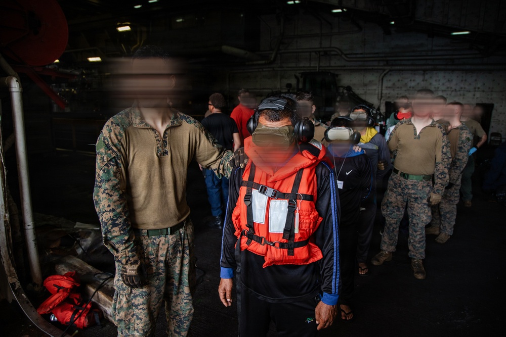 The image shows a group of individuals inside what appears to be the hangar bay or interior deck of a military vessel. Several people in U.S. Marine Corps MARPAT camouflage uniforms are present alongside civilians wearing orange life vests and hearing protection. All faces have been deliberately blurred, suggesting privacy or security considerations.