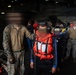 The image shows a group of individuals inside what appears to be the hangar bay or interior deck of a military vessel. Several people in U.S. Marine Corps MARPAT camouflage uniforms are present alongside civilians wearing orange life vests and hearing protection. All faces have been deliberately blurred, suggesting privacy or security considerations.