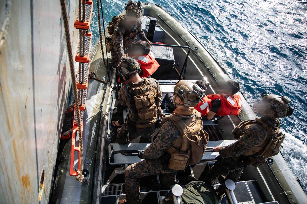 Military personnel in full tactical gear are operating from a rigid-hull inflatable boat (RHIB) alongside a larger vessel. Several individuals wearing orange life vests, distinct from the armed personnel in camouflage, are present in the boat. The scene appears to depict a maritime boarding or interdiction operation conducted at sea.