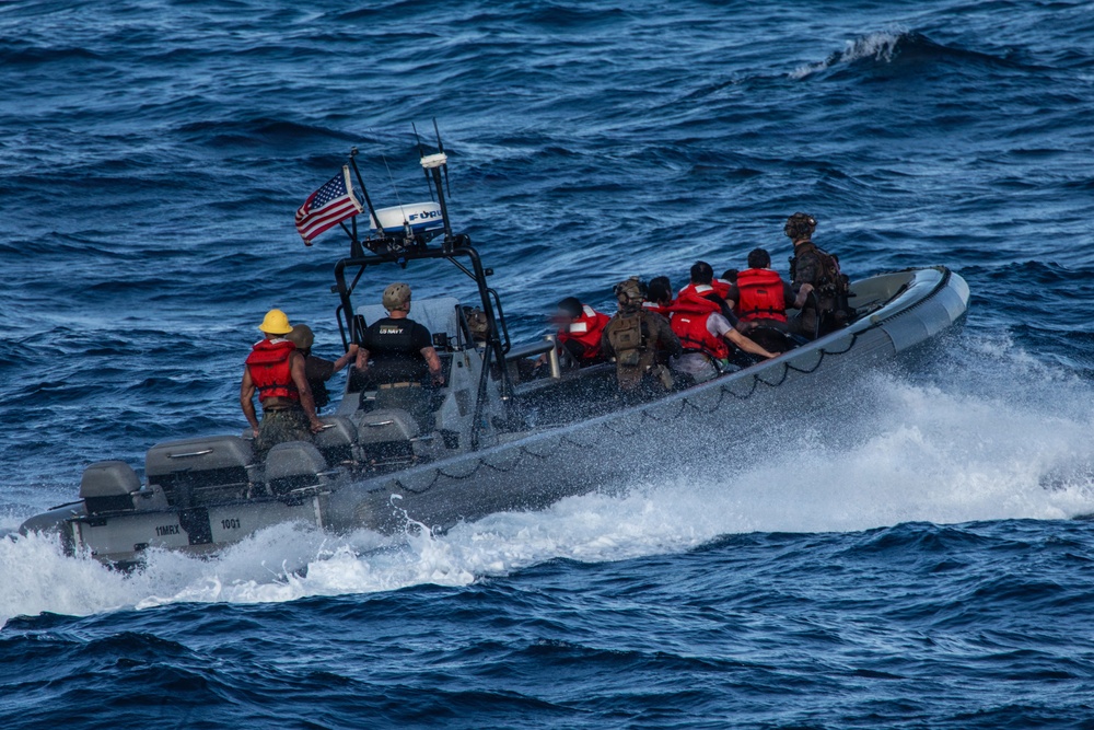 A U.S. Navy rigid-hull inflatable boat (RHIB) is underway at speed on open blue water. The vessel carries both uniformed Navy personnel and individuals wearing red life vests, suggesting a maritime interdiction or rescue operation. An American flag is visible mounted on the boat's console.