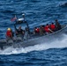 A U.S. Navy rigid-hull inflatable boat (RHIB) is underway at speed on open blue water. The vessel carries both uniformed Navy personnel and individuals wearing red life vests, suggesting a maritime interdiction or rescue operation. An American flag is visible mounted on the boat's console.