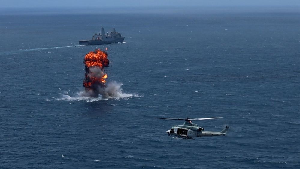 An aerial photograph captures a large explosion on the open ocean surface, with orange flames and dark smoke rising from the water. A military helicopter, identifiable as a UH-1Y Venom or similar rotary-wing aircraft, is visible in the foreground right. A large naval vessel, appearing to be an amphibious transport dock ship, is visible in the background beyond the explosion.