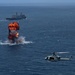 An aerial photograph captures a large explosion on the open ocean surface, with orange flames and dark smoke rising from the water. A military helicopter, identifiable as a UH-1Y Venom or similar rotary-wing aircraft, is visible in the foreground right. A large naval vessel, appearing to be an amphibious transport dock ship, is visible in the background beyond the explosion.
