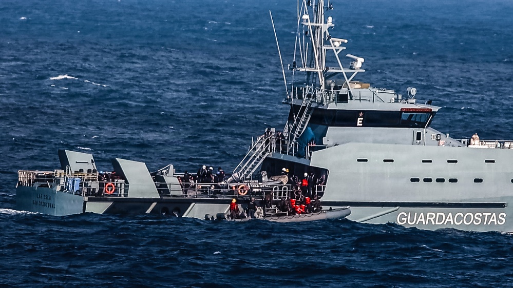 A gray coast guard vessel ('Guardacostas') is shown at sea with personnel visible on deck and a rigid inflatable boat (RIB) alongside. Multiple individuals in what appear to be life vests or rescue gear are gathered at the vessel's side, suggesting a rescue or boarding operation in progress. The sea conditions appear moderately rough with visible wave action.