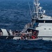 A gray coast guard vessel ('Guardacostas') is shown at sea with personnel visible on deck and a rigid inflatable boat (RIB) alongside. Multiple individuals in what appear to be life vests or rescue gear are gathered at the vessel's side, suggesting a rescue or boarding operation in progress. The sea conditions appear moderately rough with visible wave action.