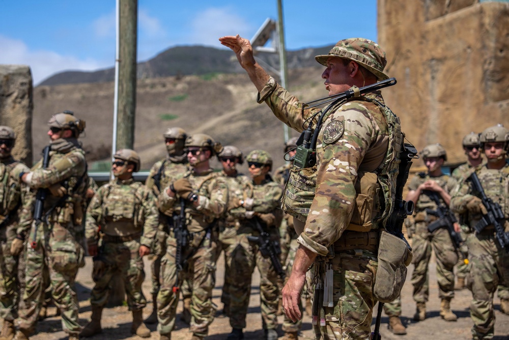 A military instructor or senior non-commissioned officer in MultiCam/OCP camouflage and a boonie hat is briefing or instructing a formation of soldiers at what appears to be a training facility. The leader is gesturing with a raised hand while addressing the assembled troops, who are equipped with rifles, body armor, and tactical gear. The setting appears to be an outdoor training range or mock urban environment with arid, hilly terrain in the background.