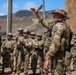 A military instructor or senior non-commissioned officer in MultiCam/OCP camouflage and a boonie hat is briefing or instructing a formation of soldiers at what appears to be a training facility. The leader is gesturing with a raised hand while addressing the assembled troops, who are equipped with rifles, body armor, and tactical gear. The setting appears to be an outdoor training range or mock urban environment with arid, hilly terrain in the background.