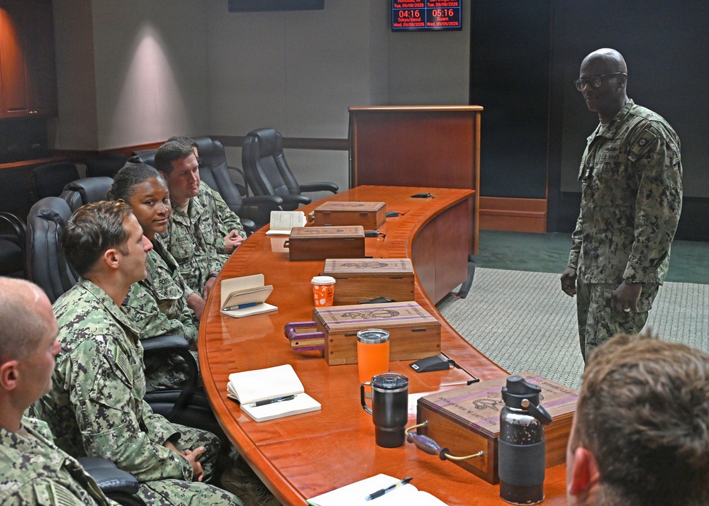 A group of U.S. Navy personnel in NWU (Navy Working Uniform) digital camouflage are gathered around a curved wooden conference table in what appears to be a command briefing room. One service member is standing and addressing the seated group, suggesting a briefing or meeting is in progress. Digital clocks on the wall display times for Tokyo/Seoul and Guam time zones, indicating a command facility with Pacific operational focus.