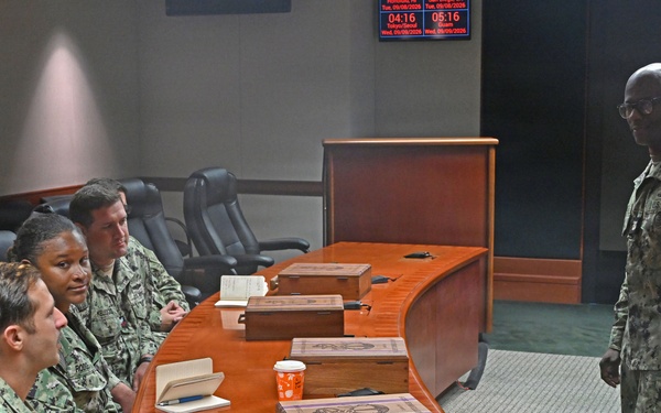 A group of U.S. Navy personnel in NWU (Navy Working Uniform) digital camouflage are gathered around a curved wooden conference table in what appears to be a command briefing room. One service member is standing and addressing the seated group, suggesting a briefing or meeting is in progress. Digital clocks on the wall display times for Tokyo/Seoul and Guam time zones, indicating a command facility with Pacific operational focus.