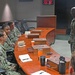 A group of U.S. Navy personnel in NWU (Navy Working Uniform) digital camouflage are gathered around a curved wooden conference table in what appears to be a command briefing room. One service member is standing and addressing the seated group, suggesting a briefing or meeting is in progress. Digital clocks on the wall display times for Tokyo/Seoul and Guam time zones, indicating a command facility with Pacific operational focus.