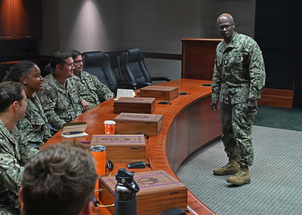 A U.S. Navy senior enlisted leader or officer is addressing a small group of Navy personnel seated around a curved wooden conference table in what appears to be a formal briefing room. The standing individual is wearing Navy Working Uniform (NWU Type III) with visible U.S. Navy tape and rank insignia. The setting suggests a leadership briefing, mentorship session, or command meeting.
