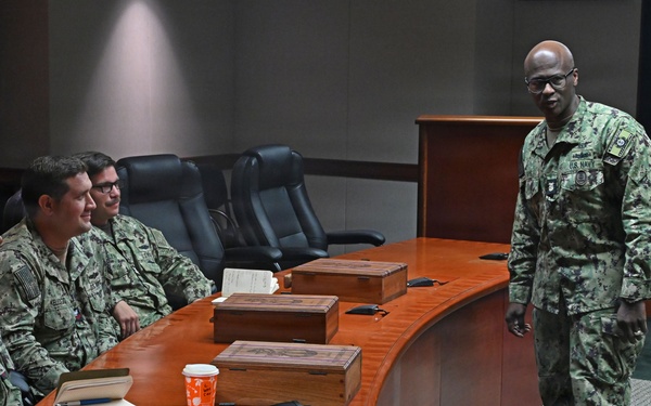 A U.S. Navy senior enlisted leader or officer is addressing a small group of Navy personnel seated around a curved wooden conference table in what appears to be a formal briefing room. The standing individual is wearing Navy Working Uniform (NWU Type III) with visible U.S. Navy tape and rank insignia. The setting suggests a leadership briefing, mentorship session, or command meeting.