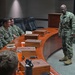 A U.S. Navy senior enlisted leader or officer is addressing a small group of Navy personnel seated around a curved wooden conference table in what appears to be a formal briefing room. The standing individual is wearing Navy Working Uniform (NWU Type III) with visible U.S. Navy tape and rank insignia. The setting suggests a leadership briefing, mentorship session, or command meeting.