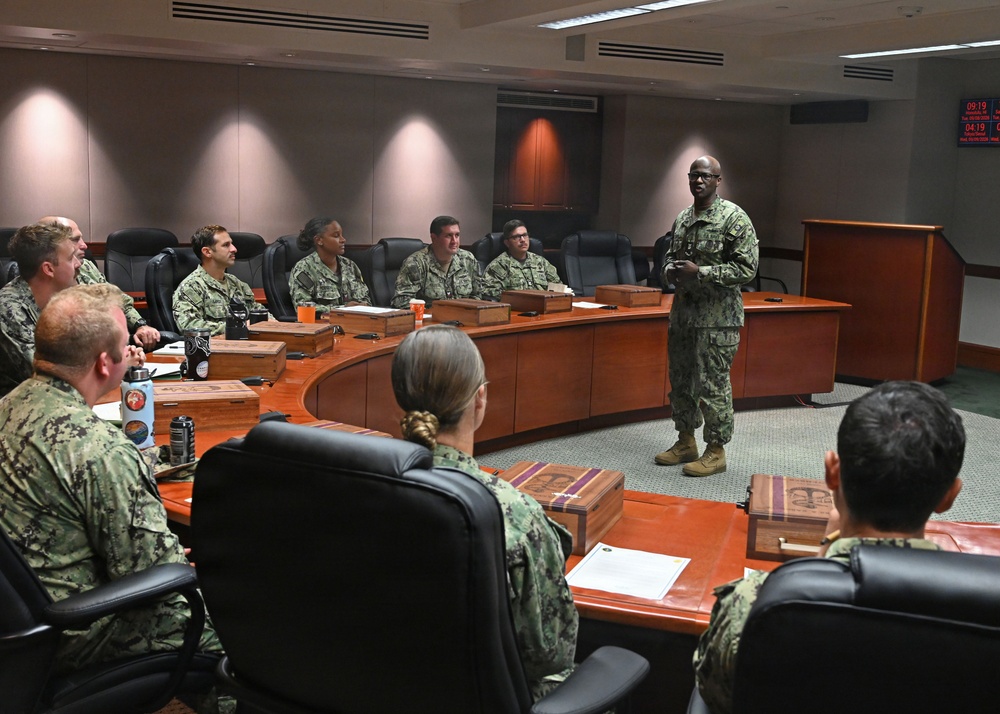 A military briefing or meeting is taking place in a formal conference room, with personnel in camouflage uniforms seated around a large curved wooden table. One individual, appearing to be a senior officer, is standing and addressing the group near a wooden podium. The setting suggests a command-level facility based on the formal furnishings and digital clock display on the wall.