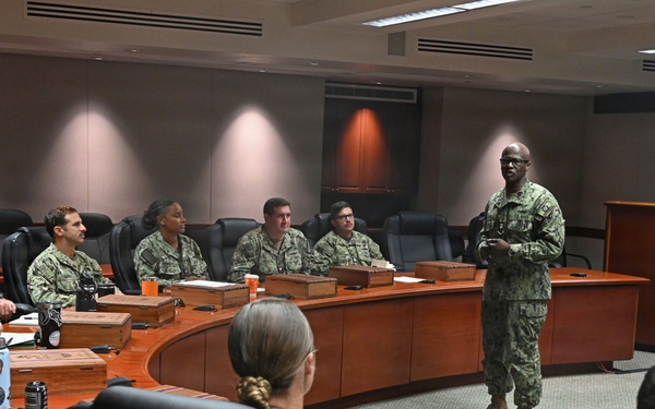 A military briefing or meeting is taking place in a formal conference room, with personnel in camouflage uniforms seated around a large curved wooden table. One individual, appearing to be a senior officer, is standing and addressing the group near a wooden podium. The setting suggests a command-level facility based on the formal furnishings and digital clock display on the wall.