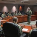 A military briefing or meeting is taking place in a formal conference room, with personnel in camouflage uniforms seated around a large curved wooden table. One individual, appearing to be a senior officer, is standing and addressing the group near a wooden podium. The setting suggests a command-level facility based on the formal furnishings and digital clock display on the wall.