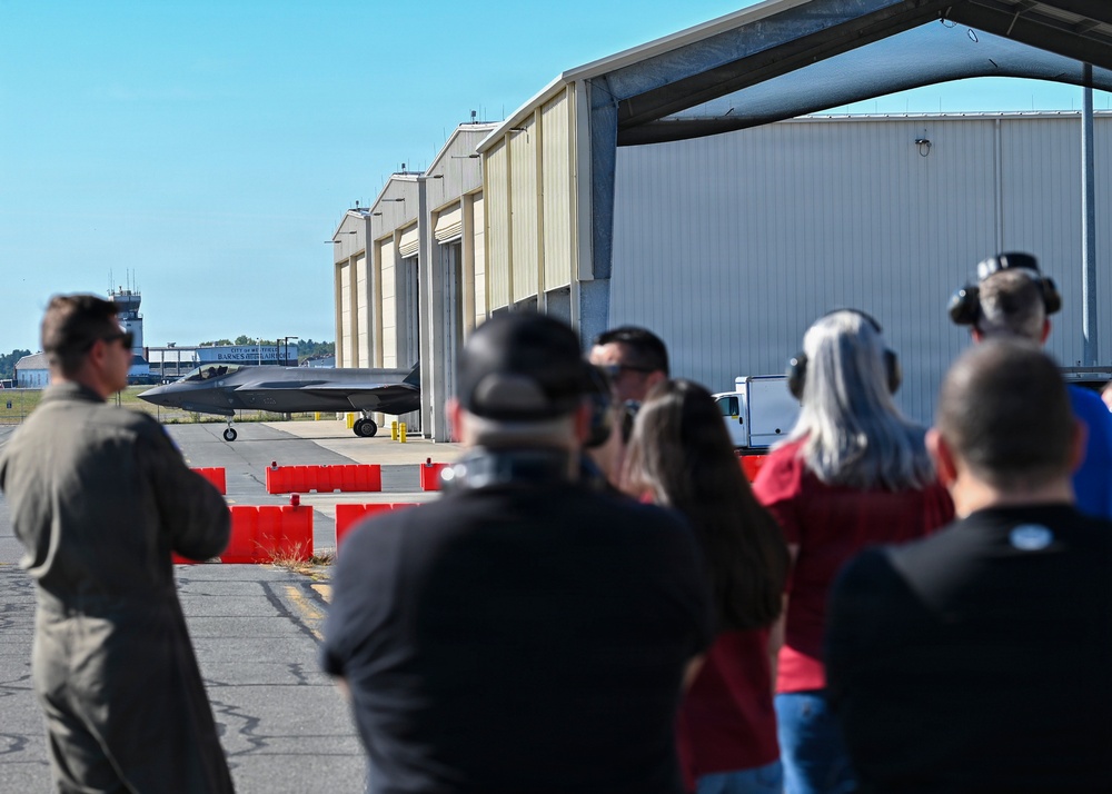 A group of spectators and a uniformed military personnel observe a stealth fighter jet (appears to be an F-22 Raptor) taxiing on a flight line near large aircraft hangars. Several observers are photographing or filming the aircraft. The setting appears to be a military air base or airshow event, with red barrier cones separating the crowd from the taxiway.