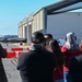 A group of spectators and a uniformed military personnel observe a stealth fighter jet (appears to be an F-22 Raptor) taxiing on a flight line near large aircraft hangars. Several observers are photographing or filming the aircraft. The setting appears to be a military air base or airshow event, with red barrier cones separating the crowd from the taxiway.