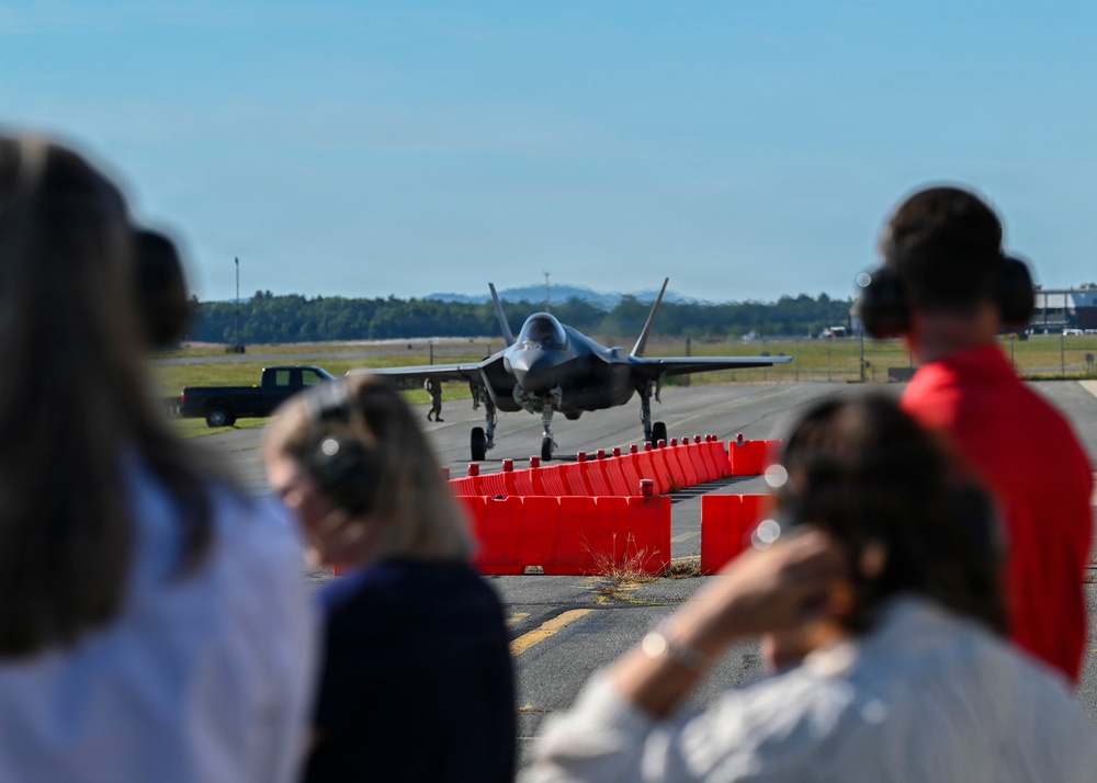 A military jet aircraft, consistent with an F-35 Lightning II, is taxiing on a runway while spectators in the foreground observe from behind a row of red safety cones/barriers. Several observers are wearing hearing protection ear muffs, suggesting a controlled viewing event or air show. The scene appears to take place at a military airbase on a clear, sunny day.