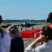 A military jet aircraft, consistent with an F-35 Lightning II, is taxiing on a runway while spectators in the foreground observe from behind a row of red safety cones/barriers. Several observers are wearing hearing protection ear muffs, suggesting a controlled viewing event or air show. The scene appears to take place at a military airbase on a clear, sunny day.