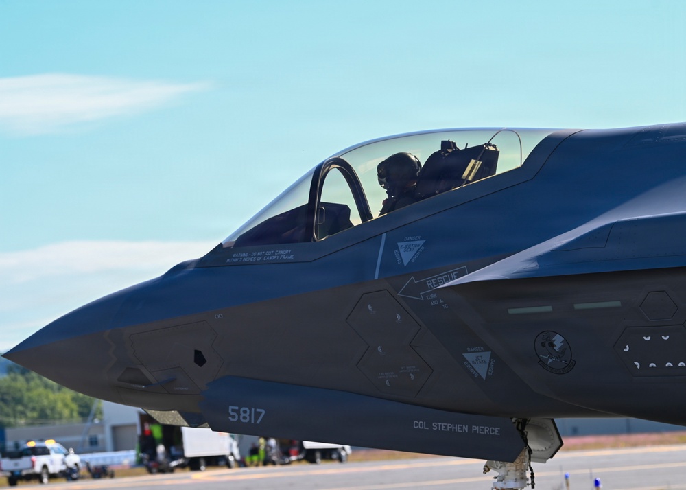 A close-up photograph of an F-35 Lightning II stealth fighter jet on a taxiway or flight line. The aircraft bears the tail number 5817 and is marked with the name 'COL STEPHEN PIERCE' below the cockpit. A pilot wearing a flight helmet is visible seated in the single-seat cockpit.