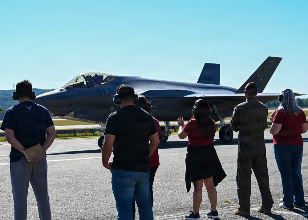 A group of observers wearing hearing protection stands on a flight line watching an F-35 Lightning II fighter jet taxi on the runway. The aircraft displays 'TX' tail codes indicating it is assigned to a Texas-based unit, with a partial tail number '5787' visible. One person in a red outfit appears to be waving at the aircraft while others observe.